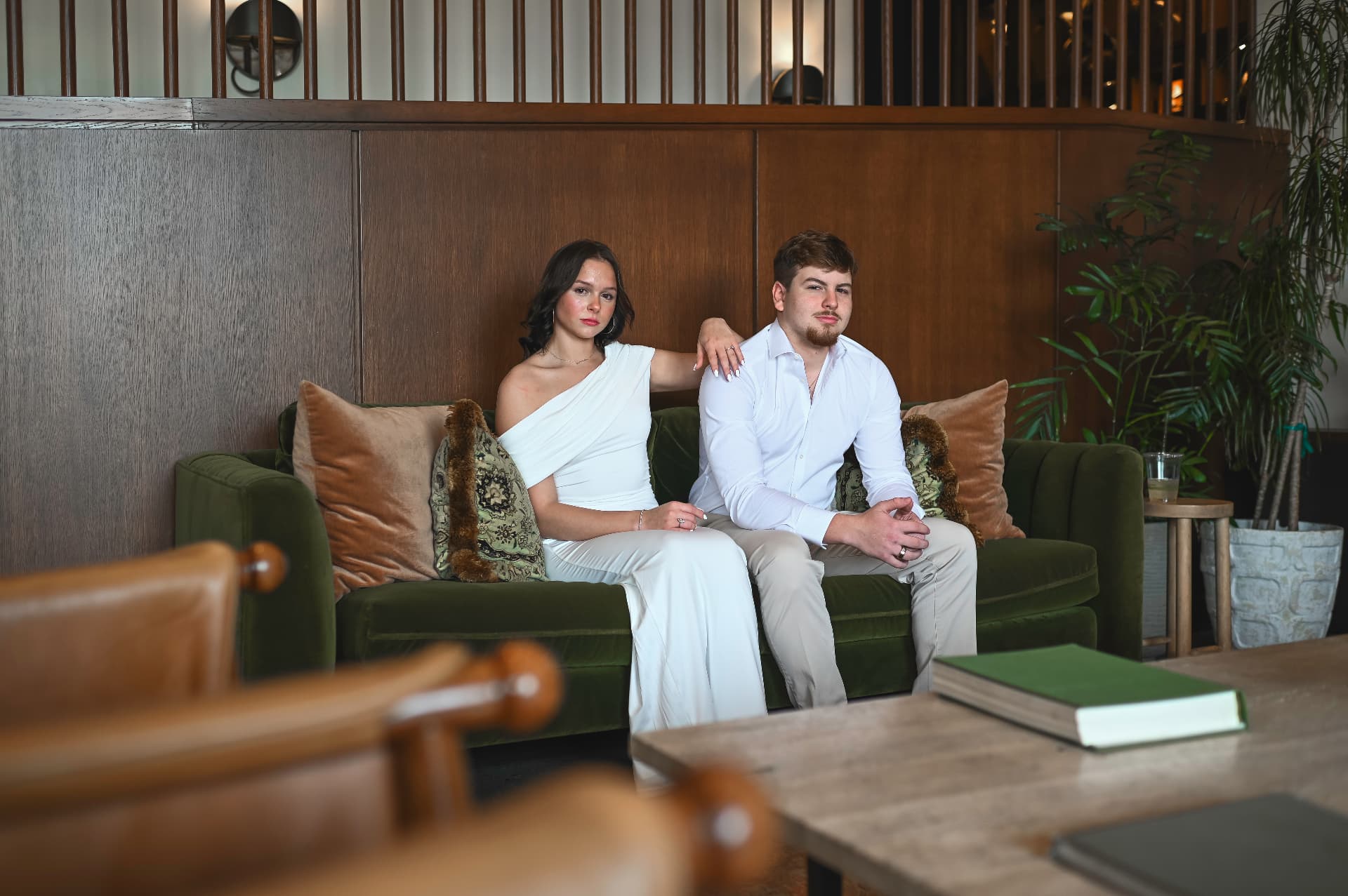 Couple in white seated on a green velvet sofa in a warm wood-paneled lounge